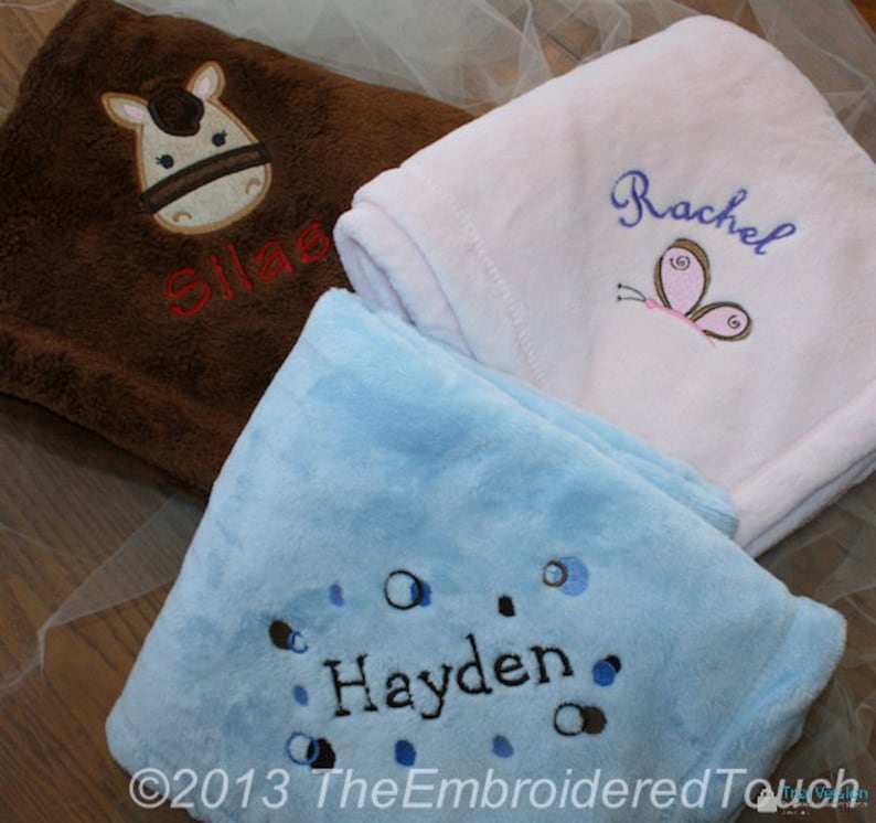 May include: Three soft, embroidered baby blankets in brown, white, and blue. The brown blanket features a horse face and the name "Silas". The white blanket has a butterfly and the name "Rachel". The blue blanket has the name "Hayden".