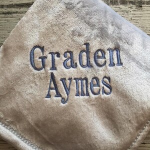 May include: A soft, gray baby blanket with the name "Graden Aymes" embroidered in blue thread. The blanket has a plush texture and a decorative stitched border. The words "The Embroidered Touch" are at the bottom.