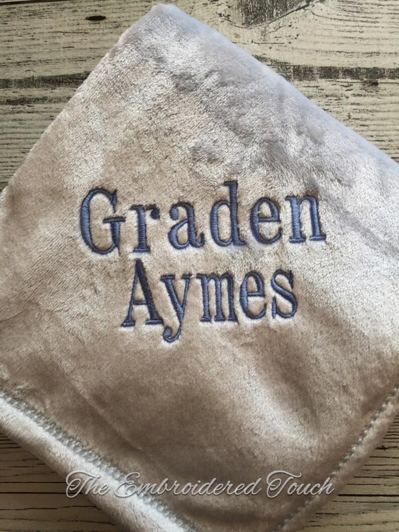 May include: A soft, gray baby blanket with the name "Graden Aymes" embroidered in blue thread. The blanket has a plush texture and a decorative stitched border. The words "The Embroidered Touch" are at the bottom.