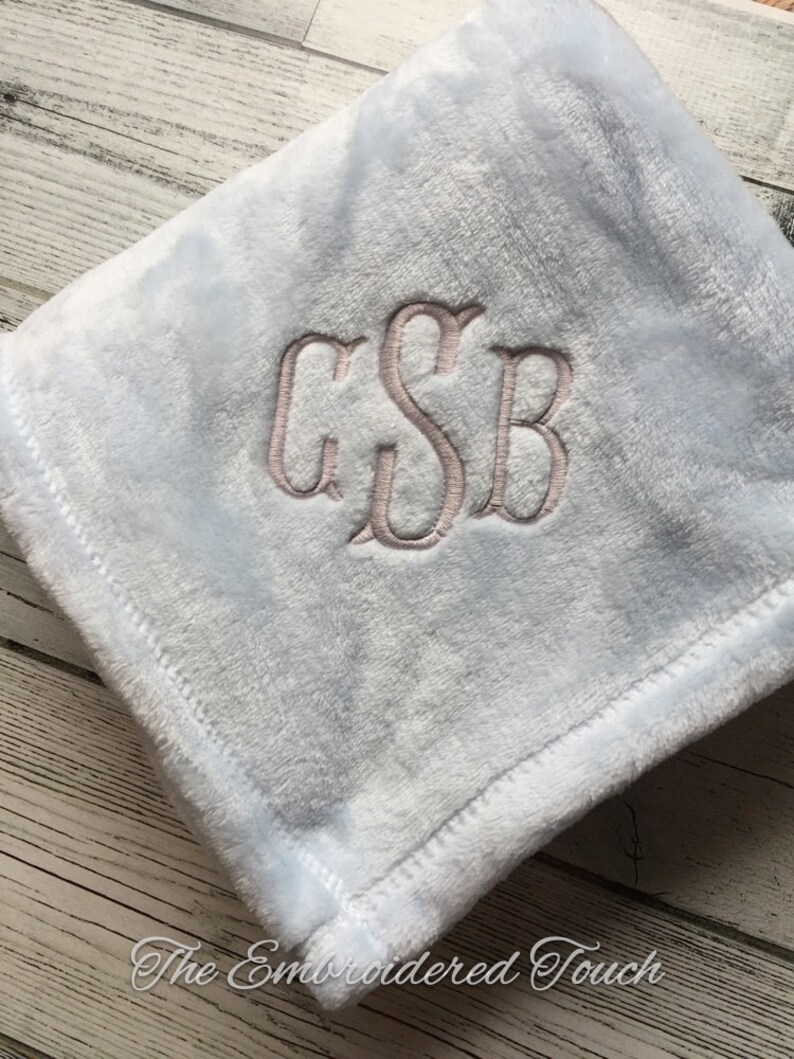 May include: A light blue plush blanket with embroidered initials "GSB" in a lighter shade. The blanket has a soft, fuzzy texture and a stitched border. The words "The Embroidered Touch" are at the bottom.