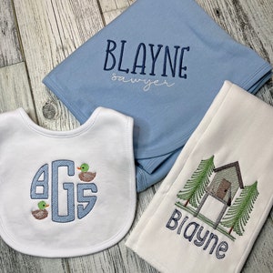 May include: A baby gift set featuring a light blue blanket embroidered with the name "Blayne Sawyer", a white bib with a monogram and ducklings, and a white burp cloth with a treehouse design and the name "Blayne".