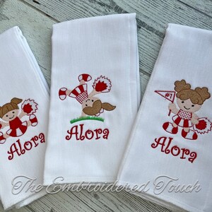 May include: Three white hand towels embroidered with cheerleader designs. Each towel features a different cheerleader pose in red, brown, and white, with the name "Alora" embroidered in red. The towels are from "The Embroidered Touch".