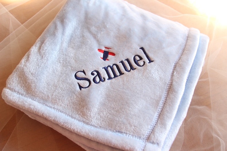 May include: A light blue baby blanket with the name "Samuel" embroidered in navy blue, along with a small red and blue airplane design. The blanket has a soft, plush texture and a stitched border.