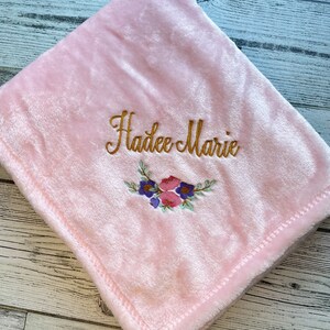 May include: A soft, pink baby blanket with the embroidered name "Hadee Marie" in gold script. A floral design in pink, purple, and blue is embroidered below the name. The blanket is folded and has a stitched edge.