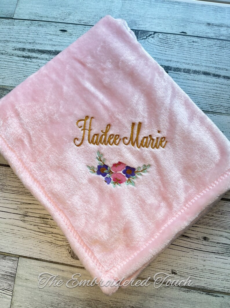 May include: A soft, pink baby blanket with the embroidered name "Hadee Marie" in gold script. A floral design in pink, purple, and blue is embroidered below the name. The blanket is folded and has a stitched edge.