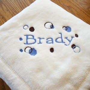 May include: A soft, cream-colored baby blanket with the name "Brady" embroidered in blue. The blanket features a pattern of blue and brown circles and is made of a plush material.