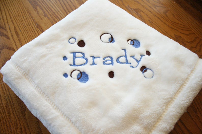 May include: A soft, cream-colored baby blanket with the name "Brady" embroidered in blue. The blanket features a pattern of blue and brown circles and is made of a plush material.