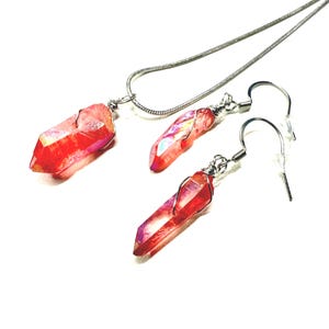 May include: A jewelry set consisting of a necklace and a pair of earrings, each featuring a raw, vibrant red crystal point wrapped in silver-toned wire. The necklace includes a thin silver-toned snake chain, while the earrings have silver-toned hooks.