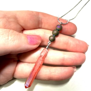 May include: A necklace featuring a long, translucent pink crystal pendant wrapped in silver wire, connected to three small, spherical, iridescent dark green beads. The pendant hangs from a thin, silver-toned metal chain.