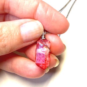 May include: A pendant featuring a raw, faceted crystal with vibrant pink and red hues, secured by a silver-toned wire wrap at the top. The crystal is attached to a thin, metallic silver-colored chain, showcasing a natural, angular shape.