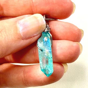 May include: A pendant featuring a translucent, iridescent blue-green crystal point wrapped in silver-toned wire. The crystal has a faceted, elongated shape with a shimmering surface that reflects light, suspended from a thin, metallic chain.