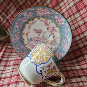 Op de afbeelding: Een wit porseleinen theekopje met een bloemenmotief in tinten blauw, geel en roze. Het kopje heeft een gouden rand en staat op een rood-wit geruit tafelkleed.