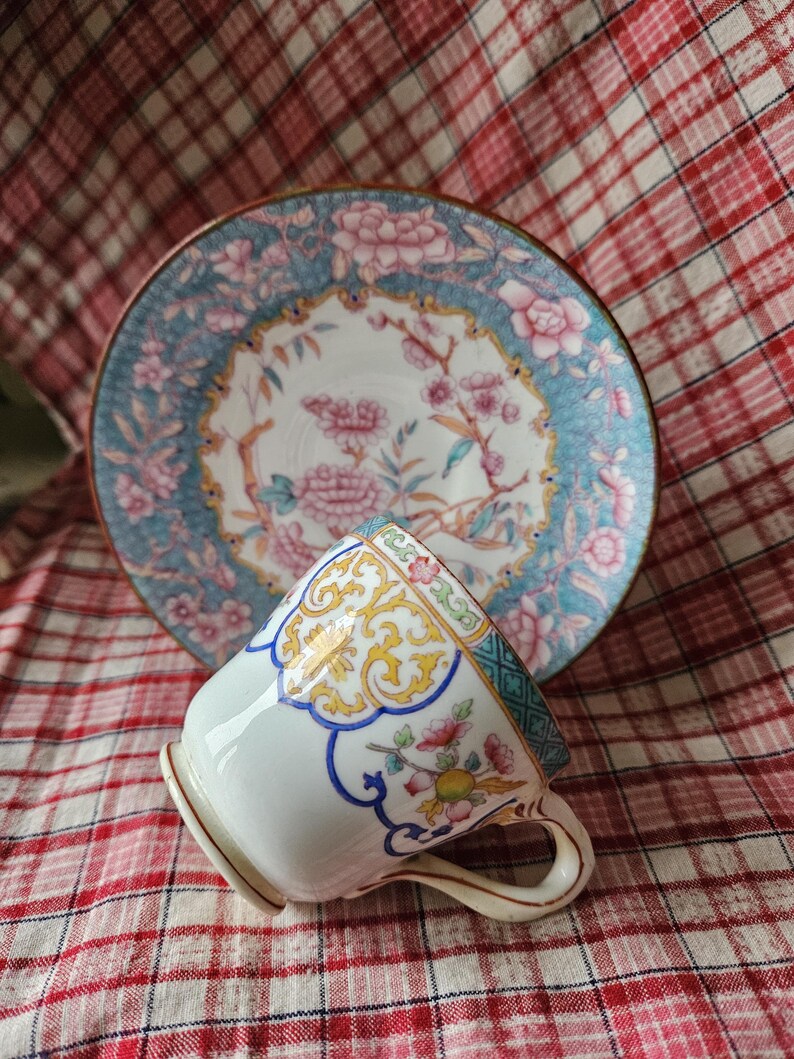 Op de afbeelding: Een wit porseleinen theekopje met een bloemenmotief in tinten blauw, geel en roze. Het kopje heeft een gouden rand en staat op een rood-wit geruit tafelkleed.