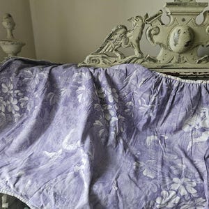 Puede incluir: Una tela lavanda drapeada con un estampado floral y de figuras en blanco. La tela cubre un cabecero decorativo blanco roto envejecido con detalles tallados. El cabecero presenta motivos de aves y flores.