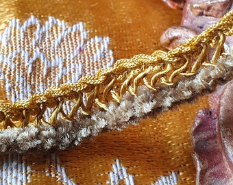 May include: Detailed shot of gold decorative trim with a fringed edge. The trim is set against a yellow and white patterned fabric, creating a contrast of textures and colours. The trim's design adds a touch of elegance.