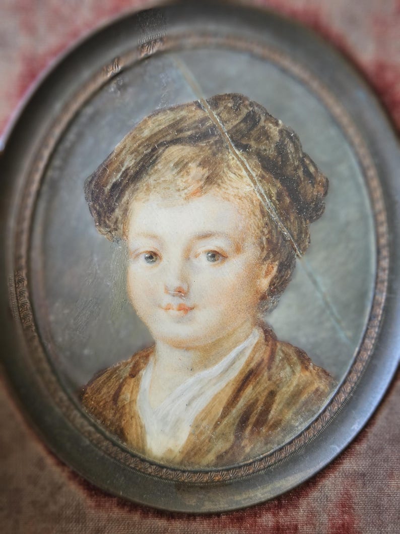 May include: Oval portrait miniature of a young person with light skin, brown hair, and a brown jacket. The portrait is set in a decorative, oval metal frame. The painting has a soft, antique aesthetic.