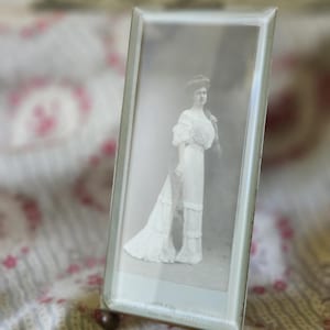 May include: A rectangular glass picture frame with a beveled edge and two small metal ball feet. The frame holds a vintage black and white photograph of a person standing in a long, light-colored dress with ruffled details and a dark stole.