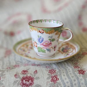 Op de afbeelding: Een witte porseleinen theekop en schotel set met een delicaat handgeschilderd bloemmotief in roze, blauwe en groene tinten. De rand van de kop en schotel is versierd met een groen en goudkleurige patroonrand en de kop heeft een gebogen oortje.