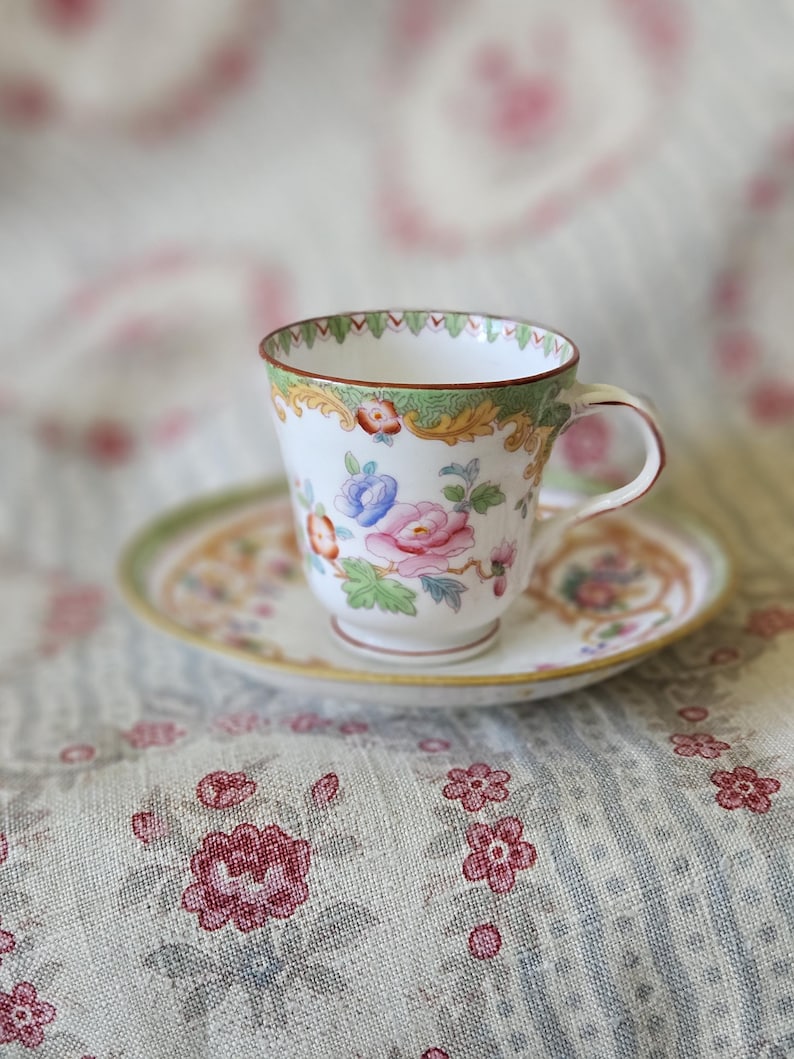 Op de afbeelding: Een witte porseleinen theekop en schotel set met een delicaat handgeschilderd bloemmotief in roze, blauwe en groene tinten. De rand van de kop en schotel is versierd met een groen en goudkleurige patroonrand en de kop heeft een gebogen oortje.