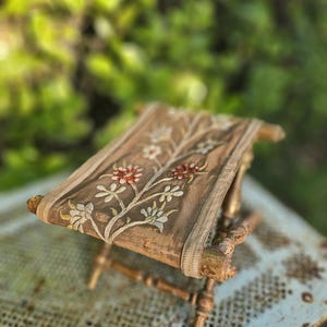 Può includere: Uno sgabello pieghevole di piccole dimensioni con seduta in tessuto marrone decorata da motivi floreali ricamati in rosso, bianco e verde. La struttura è realizzata in legno marrone chiaro usurato, con gambe tornite e rinforzo a croce.