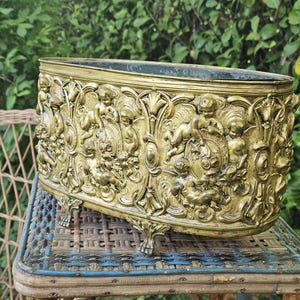 Puede incluir: Una jardinera antigua dorada con detalles de relieve ornamentados. La jardinera rectangular presenta figuras de querubines y diseños florales. Descansa sobre cuatro patas decorativas y está colocada sobre una mesa de mimbre azul y marrón.