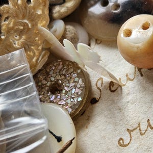 May include: A collection of vintage buttons, including a white button with iridescent glitter, a brown button with four holes, and a white button with a black thread attached. The buttons are arranged on a piece of paper with handwritten text.