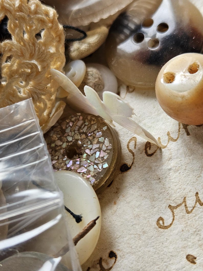 May include: A collection of vintage buttons, including a white button with iridescent glitter, a brown button with four holes, and a white button with a black thread attached. The buttons are arranged on a piece of paper with handwritten text.