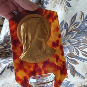 Puede incluir: Una placa de pared con estampado de caparazón de tortuga con una imagen dorada de la Virgen María y un pequeño recipiente de vidrio.