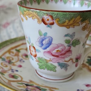 Op de afbeelding: Een witte porseleinen theekop en schotel met een handgeschilderd bloemmotief in roze, blauwe en oranje tinten. De rand is versierd met een groene en gouden krulrand en de onderkant van de kop heeft een kleine voet.