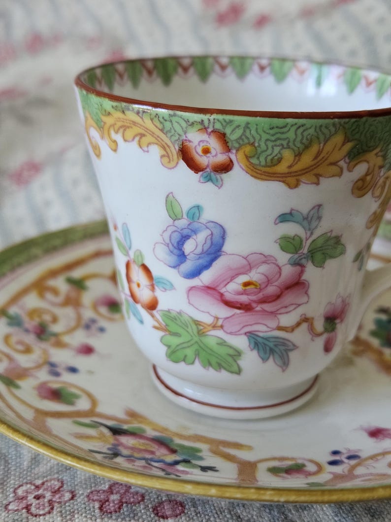 Op de afbeelding: Een witte porseleinen theekop en schotel met een handgeschilderd bloemmotief in roze, blauwe en oranje tinten. De rand is versierd met een groene en gouden krulrand en de onderkant van de kop heeft een kleine voet.