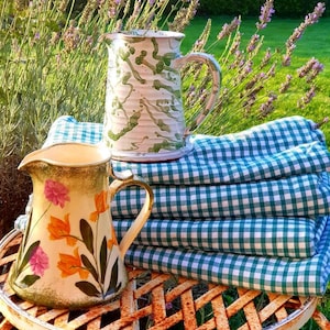 Puede incluir: Dos jarras de cerámica con diseños florales, una verde y blanca, la otra con flores naranjas y rosas, sobre un mantel a cuadros verde y blanco en una mesa de metal.