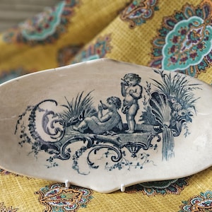 Może przedstawiać: Owalny, kremowy ceramiczny talerz z niebiesko-białym wzorem transferowym. Wzór przedstawia dwa cherubiny, liście i muszlę. Talerz jest umieszczony na żółtej tkaninie z wzorem paisley.