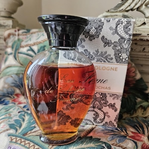 May include: A vintage bottle of "Eau de Cologne Femme" by Marcel Rochas. The bottle is clear glass with a black cap and a round, bulbous shape. The liquid inside is a deep amber colour. The bottle is sitting on a floral patterned fabric.