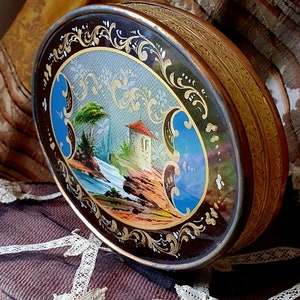 May include: An oval-shaped, antique tin box with a gold-colored rim and a painted scene of a small house and trees on a rocky shore. The scene is surrounded by a black and gold floral design.