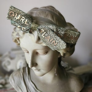 May include: A marble bust of a woman wearing a glittery bow with the words "GURL" and "PLAIN" in gold lettering. The bow is silver and green. The bust is a classical sculpture.