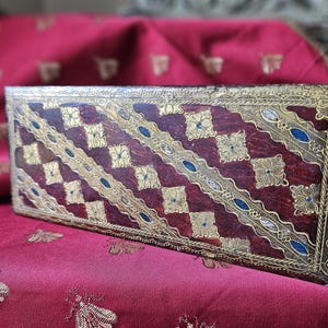 May include: Rectangular wooden box with a dark red background and gold and blue decorative accents. The box features a repeating pattern of gold floral squares and diagonal gold lines with blue oval details.