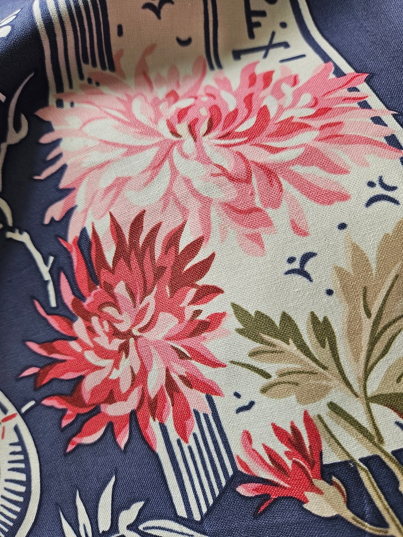 May include: Close-up of a fabric with a floral pattern. The design features large pink and red chrysanthemum flowers, green leaves, and white accents on a navy blue background. The fabric has a vintage or retro aesthetic.