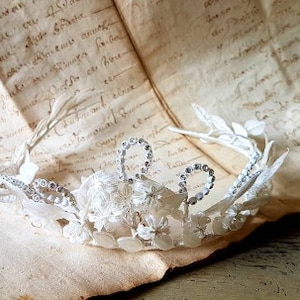 May include: A white floral crown with a delicate design and silver accents. The crown is made of lace and beads and has a vintage look.