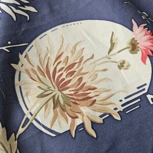 May include: Close-up of a blue fabric with a floral pattern. The design features a large, stylized flower in shades of brown, beige, and pink, set within a white oval. Additional white floral elements and geometric shapes are scattered across the fabric.