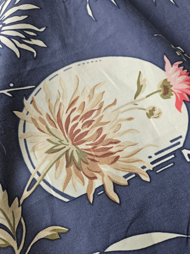 May include: Close-up of a blue fabric with a floral pattern. The design features a large, stylized flower in shades of brown, beige, and pink, set within a white oval. Additional white floral elements and geometric shapes are scattered across the fabric.