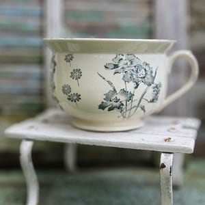 May include: A cream-coloured ceramic chamber pot featuring a dark grey floral pattern of various blossoms and stems. The vessel has a wide rim, a curved handle on the right side, and a circular base, resting on a small white metal stand.