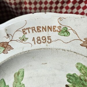 May include: A white ceramic plate with a brown and green floral design. The plate has the words "ETRENNES 1895" written in gold lettering.