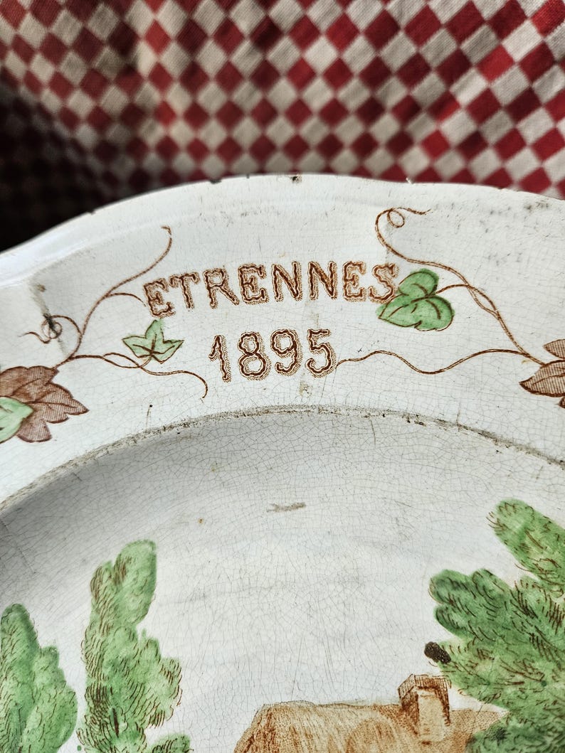 May include: A white ceramic plate with a brown and green floral design. The plate has the words "ETRENNES 1895" written in gold lettering.