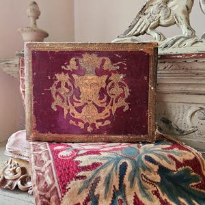 May include: A small, red, leather-bound box with gold accents and a decorative design featuring a stylized face and floral motifs.