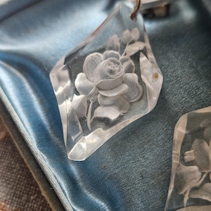 May include: A pair of clear, diamond-shaped earrings featuring an intricate, frosted intaglio carving of a single rose with leaves. The earrings are suspended from a metal finding and are presented within a blue, satin-lined jewelry box.