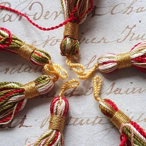 May include: Six colorful tassels with gold accents. The tassels are made of red, white, green, and gold thread. They are arranged in a circle on a white surface with handwritten text.