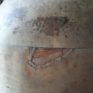 May include: A vintage wooden hat form with a crack in the side. The form is made of light brown wood and has a smooth surface. The crack is visible on the side of the form and is filled with a dark brown material.