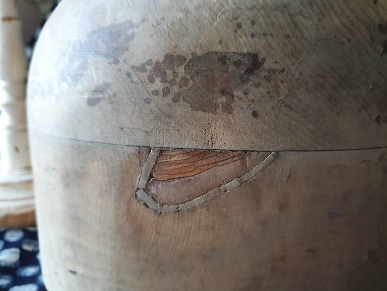May include: A vintage wooden hat form with a crack in the side. The form is made of light brown wood and has a smooth surface. The crack is visible on the side of the form and is filled with a dark brown material.