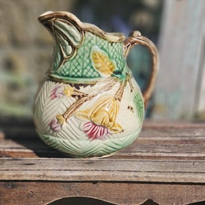 Puede incluir: Una jarra de cerámica con un patrón de celosía texturizado en relieve de color verde claro. El cuerpo está decorado con motivos florales en relieve en amarillo, rosa y marrón, con un asa esmaltada en marrón y un borde festoneado con detalles marrones.