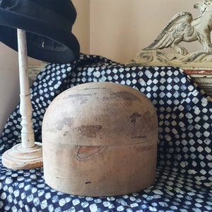 May include: A wooden hat block with a worn, natural finish. The block is round and has a slight indentation at the top. It is sitting on a blue and white patterned fabric.